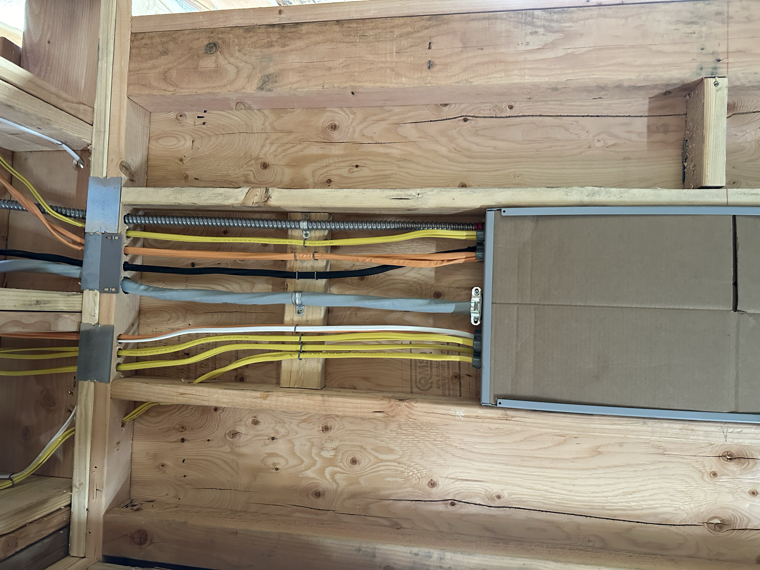 Interior view of a wooden framed wall with electrical wiring, conduits, and a metal junction box in a building under construction.