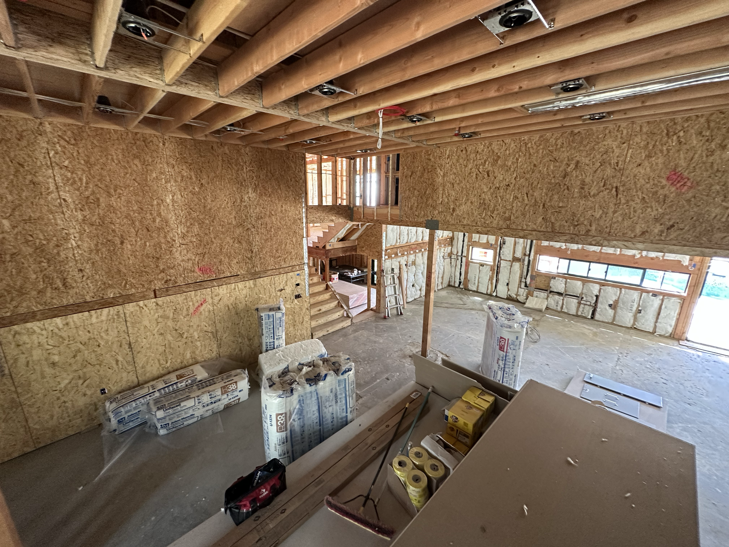 Interior view of a house under construction with exposed wooden framing, insulation, stairs, and construction materials scattered on the floor.