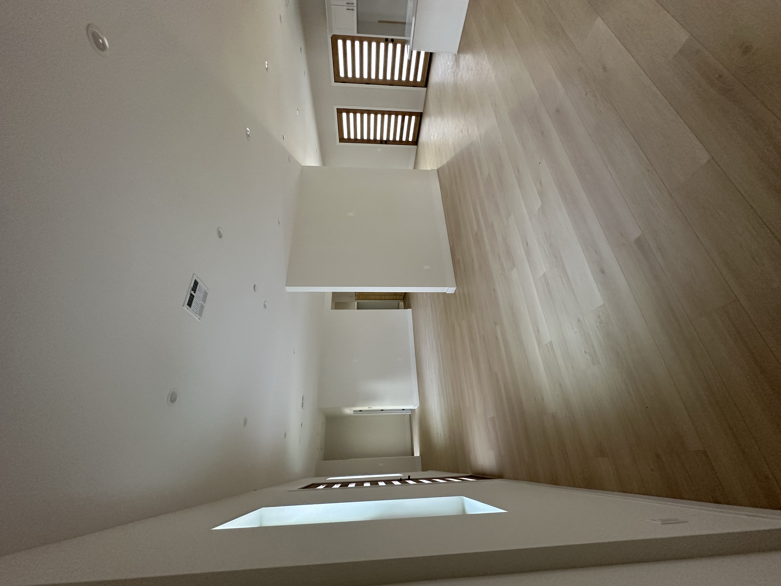 An empty living room with hardwood flooring, white walls, and a white ceiling with recessed lighting. There is a window with wooden slats in the background.