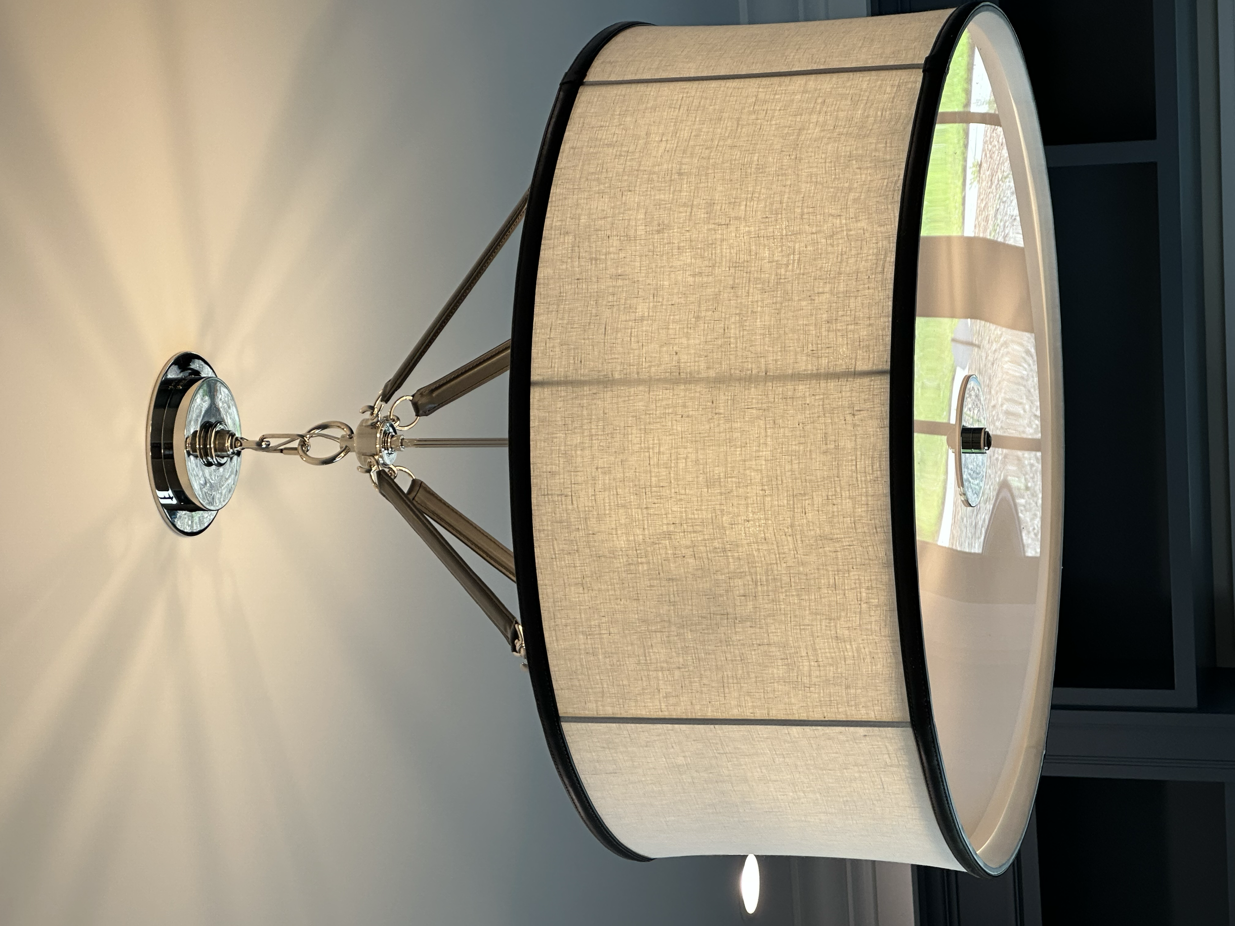 Close-up view of a modern table lamp with a beige fabric shade and metal fixtures, placed on a white surface near a window with a view of green grass outside.