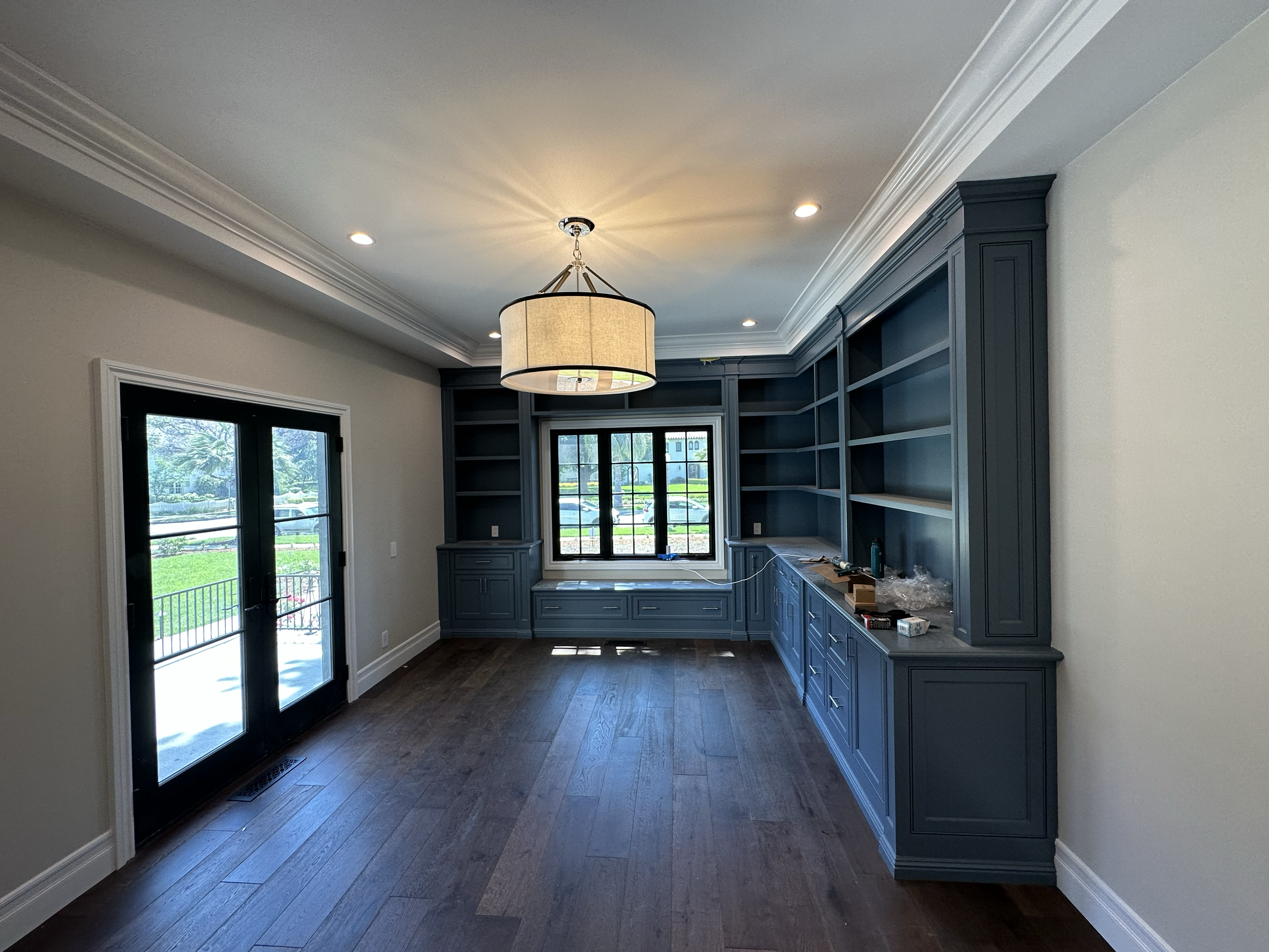 Living room with dark blue built-in bookshelves and cabinets, two large windows, wooden flooring, and a ceiling light fixture, with construction tools and materials on the countertop.