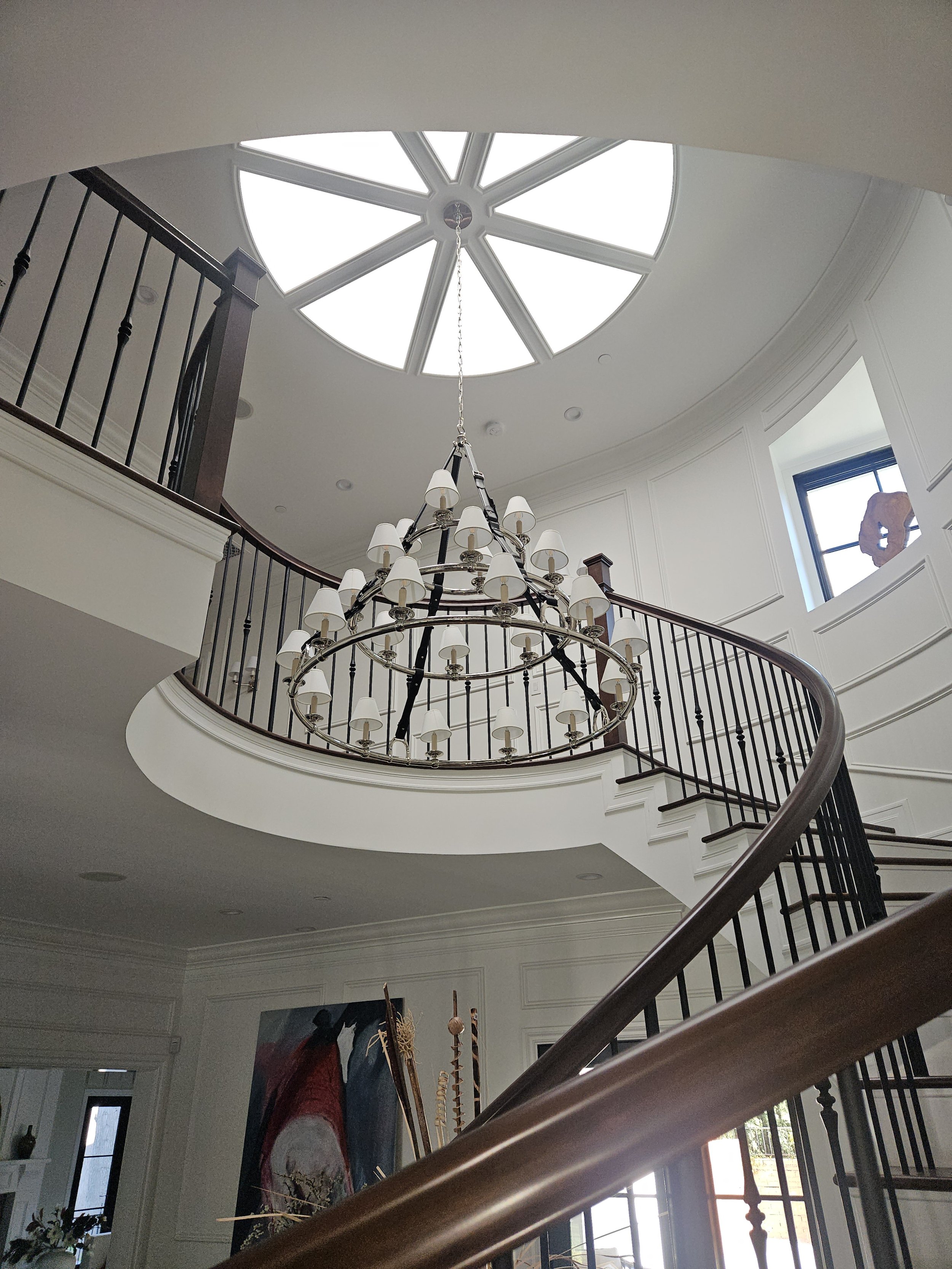 Interior view of a large, elegant staircase with a chandelier hanging above, facing upward towards a skylight with a circular, segmented design.