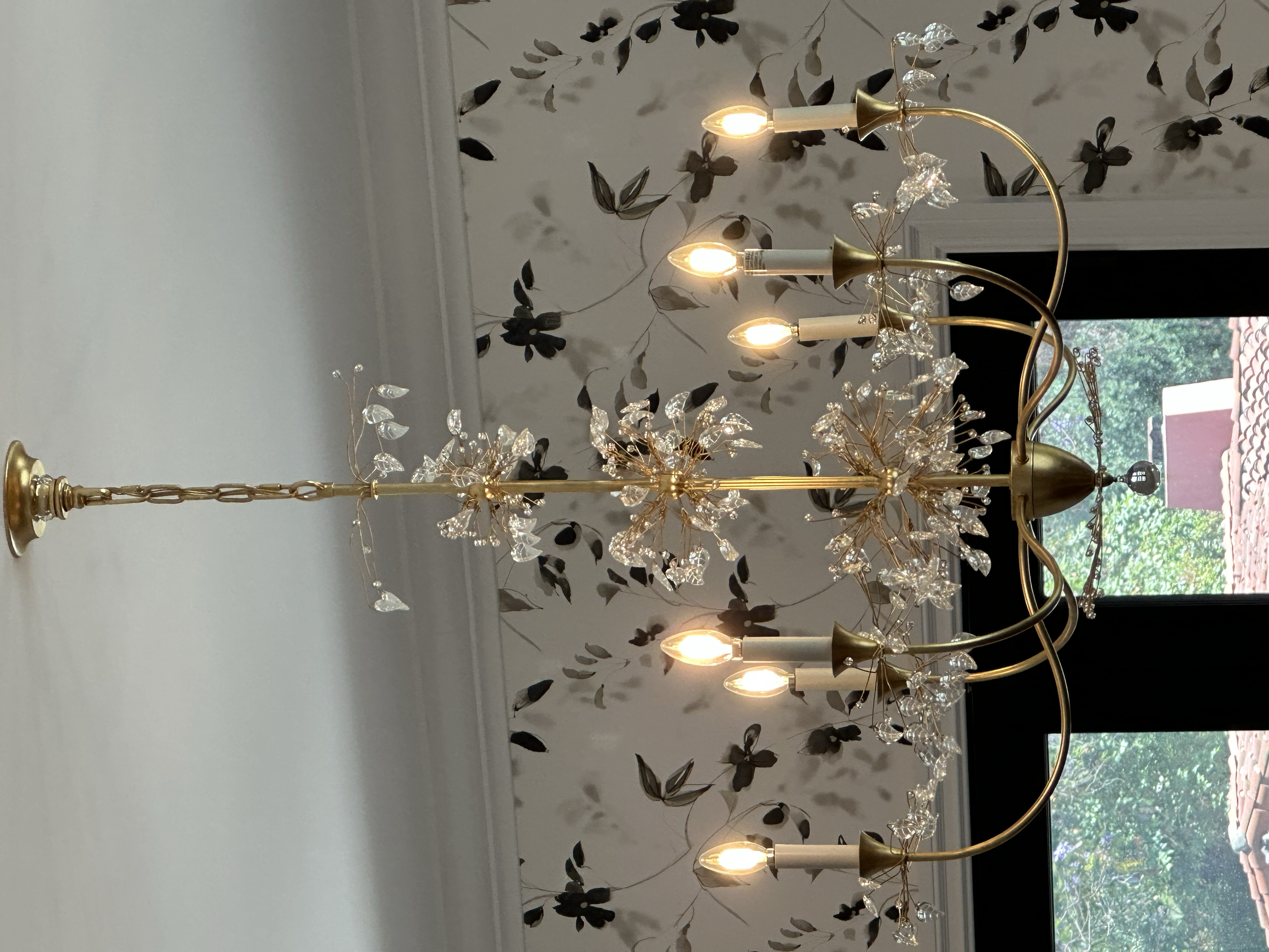 A chandelier with multiple candle-shaped lights and decorative crystal-like leaves hangs from a ceiling. In the background, there is a window with a view of trees and a building with a tiled roof.