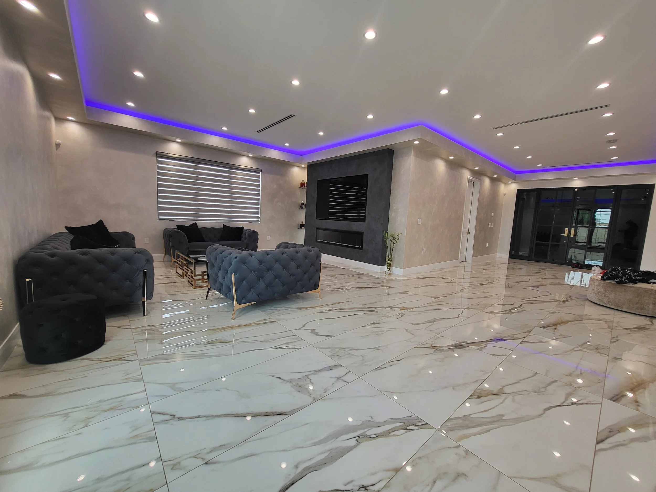 A modern living room with white marble flooring, a wall-mounted flat-screen TV, and comfortable grey sofas arranged around a glass coffee table. The ceiling has recessed lighting and purple LED accent lights, with a window with horizontal blinds on t