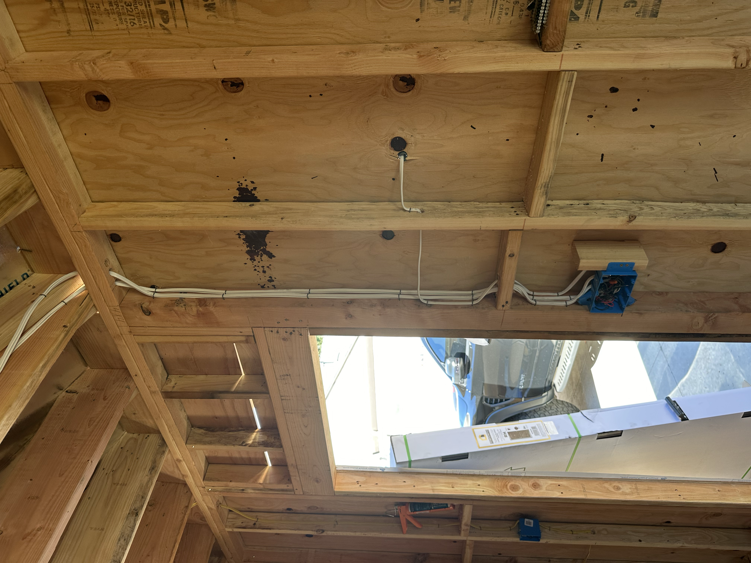 Interior view of a wooden framing structure in a building under construction, showing electrical wiring, outlets, and open wall spaces, with a window and garage visible outside.