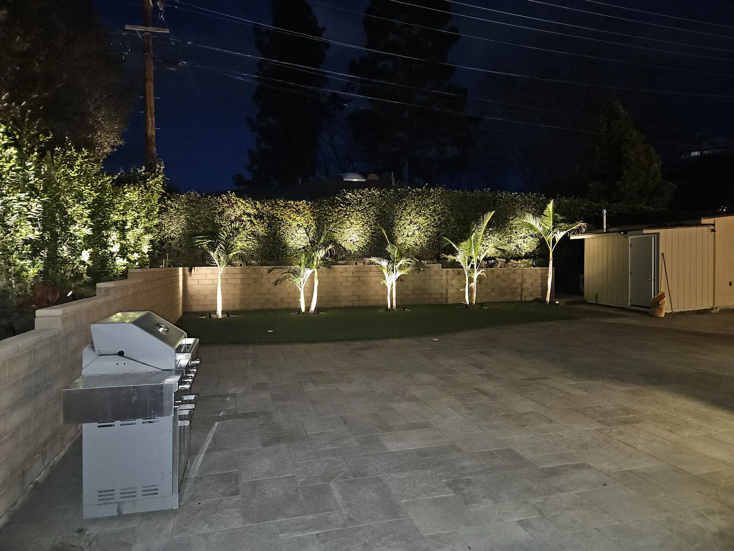 An outdoor patio area at night with lit palm trees, a beige brick wall, grills, and a storage shed. Trees and power lines are visible in the background.