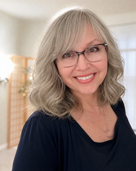 A smiling woman with shoulder-length gray hair and glasses, wearing a navy blue top and a silver necklace, standing indoors in a well-lit room.