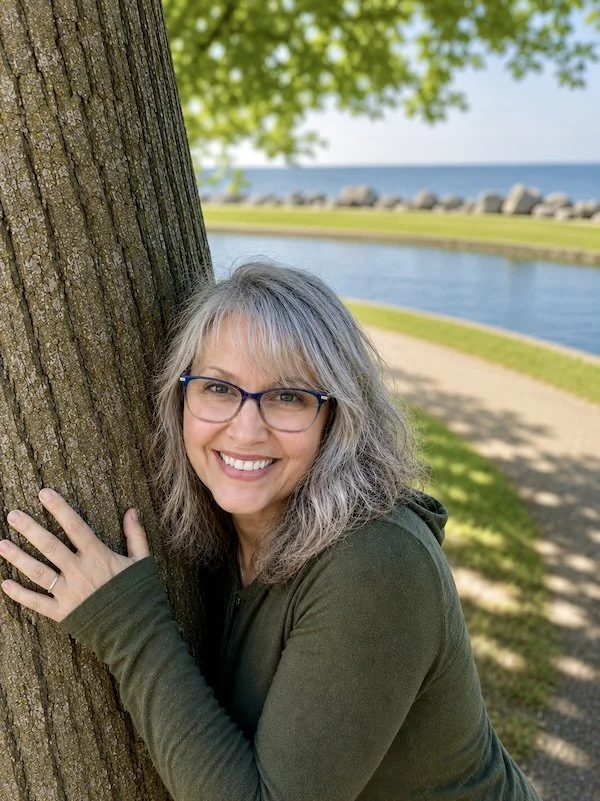 Kim smiling and hugging a tree near a lake, ready to share about herself and her yoga therapy and wellness studio.