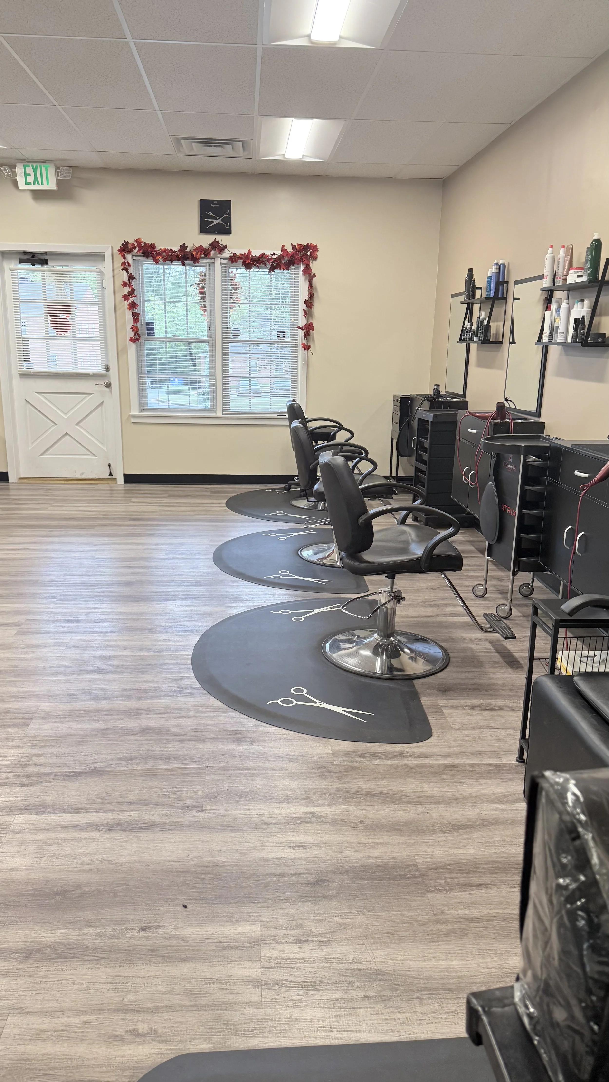 An empty hair salon with four styling chairs on circular mats with scissors and comb illustrations, shelves with hair products, a mirror, windows with red leaf decorations, and a door with a window and blinds.
