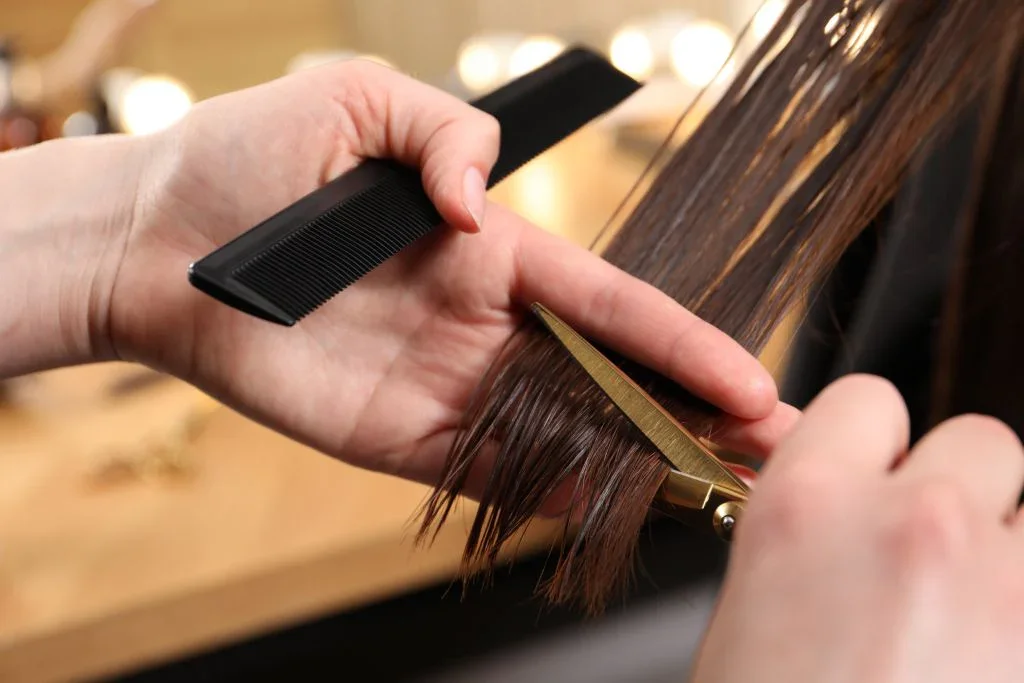 A person is cutting a client's brown hair with scissors while holding a black comb.