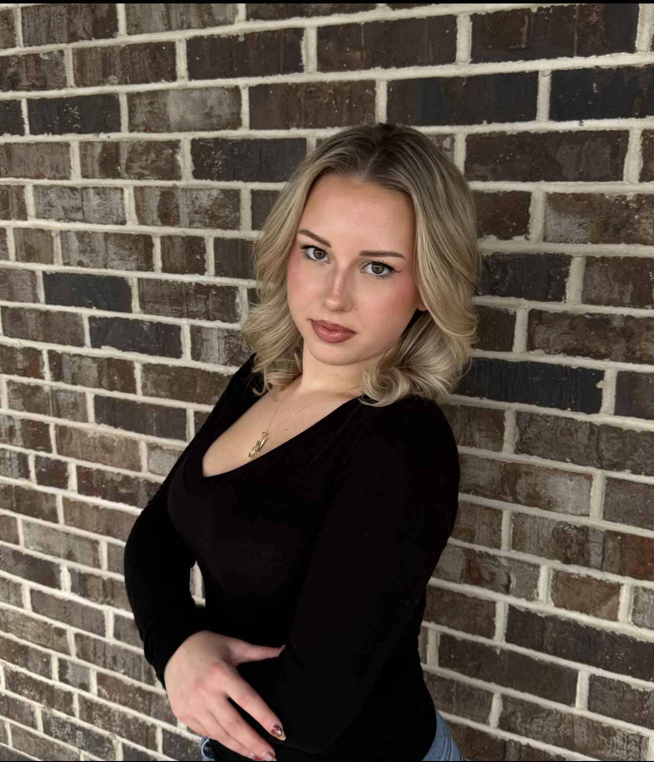 A young woman with blonde hair styled in soft curls, wearing a black top, standing against a brick wall with her arms crossed, looking at the camera.