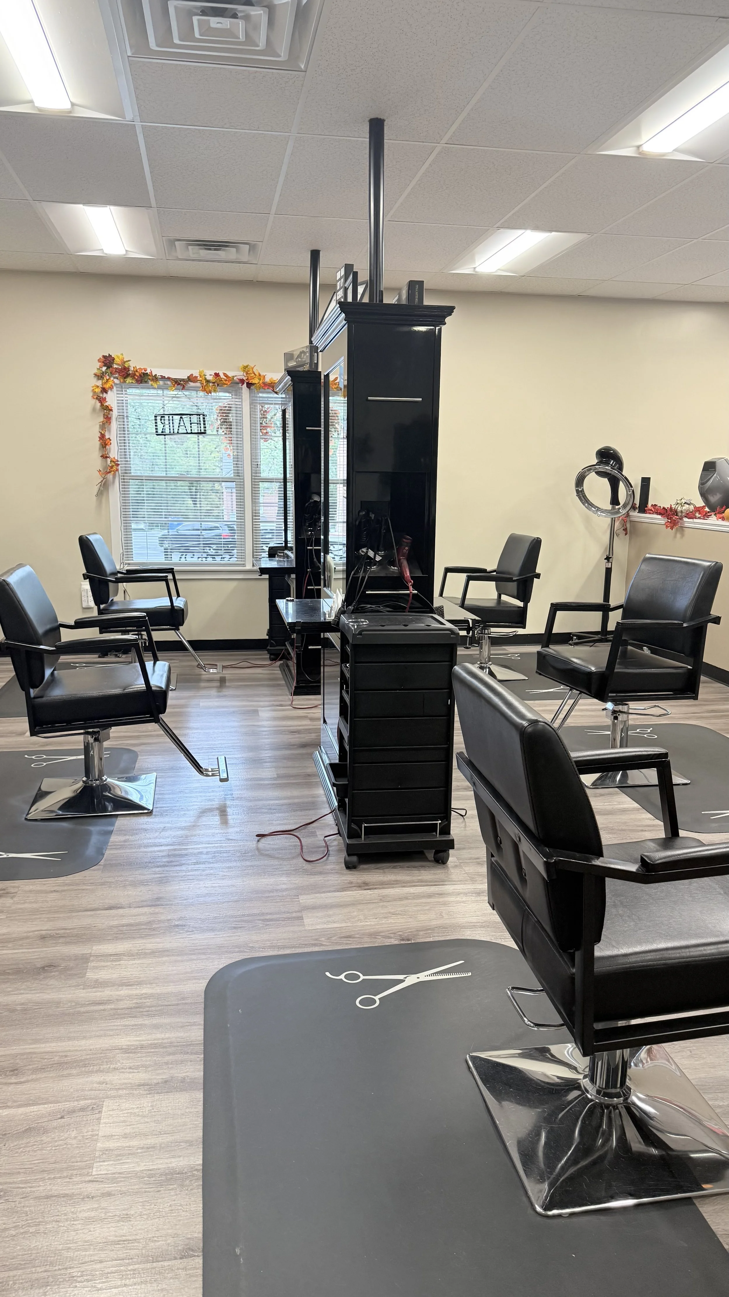 Empty hair salon with four black styling chairs, mirrors, and scissors markings on mats on the light wood floor. Halloween decorations with orange and red leaves around the window, and a hair steamer on the right side.