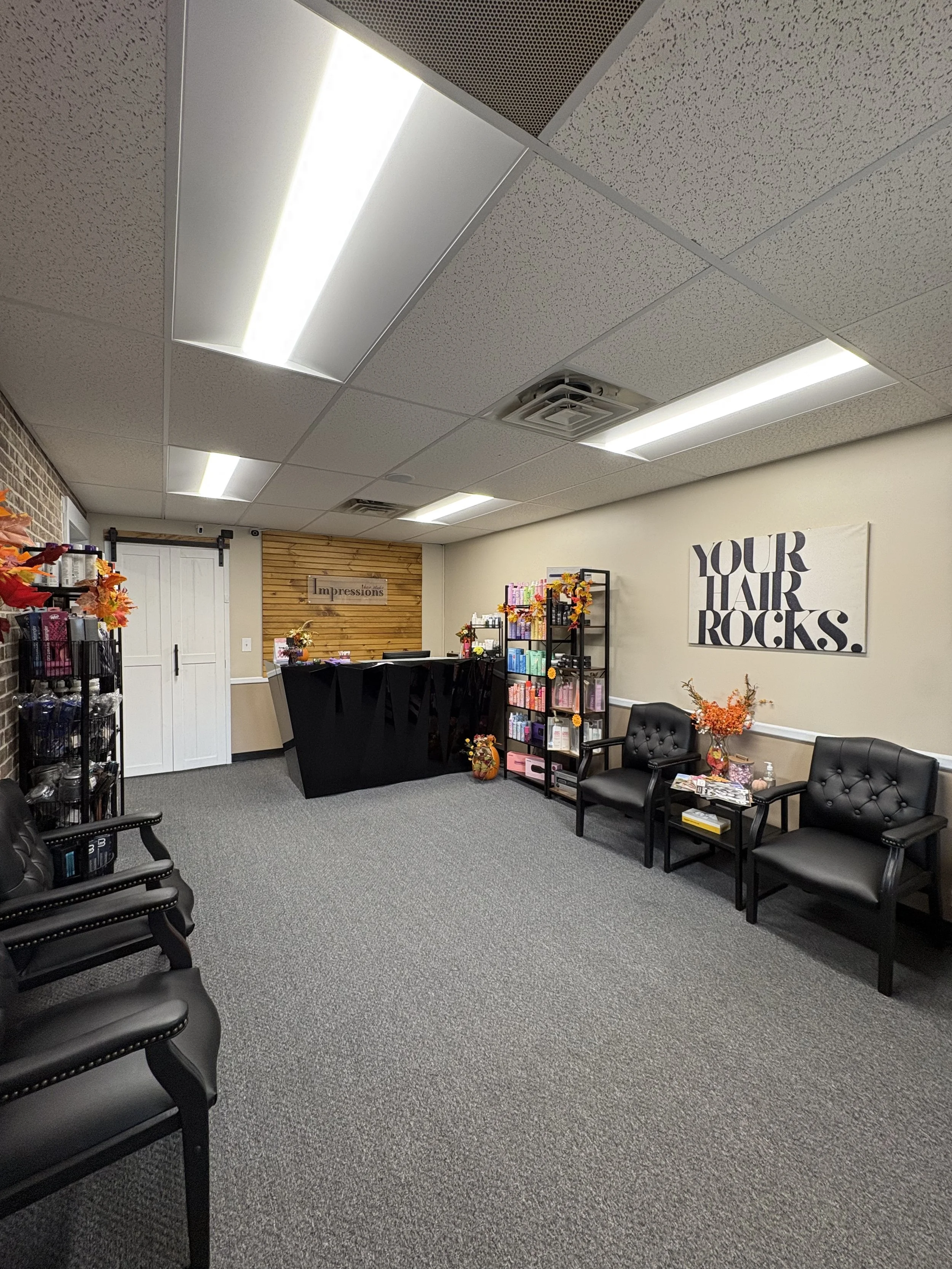 An indoor waiting area with black cushioned chairs, some with armrests, on a gray carpet. The wall on the right has a large sign that says "Your Hair Rocks" and shelving with colorful products. The back wall has a sign that says "Impressions." The ceiling has fluorescent lights and ventilation vents.