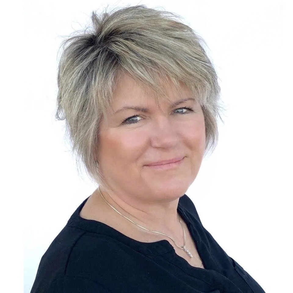 A middle-aged woman with short blonde hair, blue eyes, and light makeup, smiling slightly at the camera, wearing a black top and a silver necklace, against a plain white background.