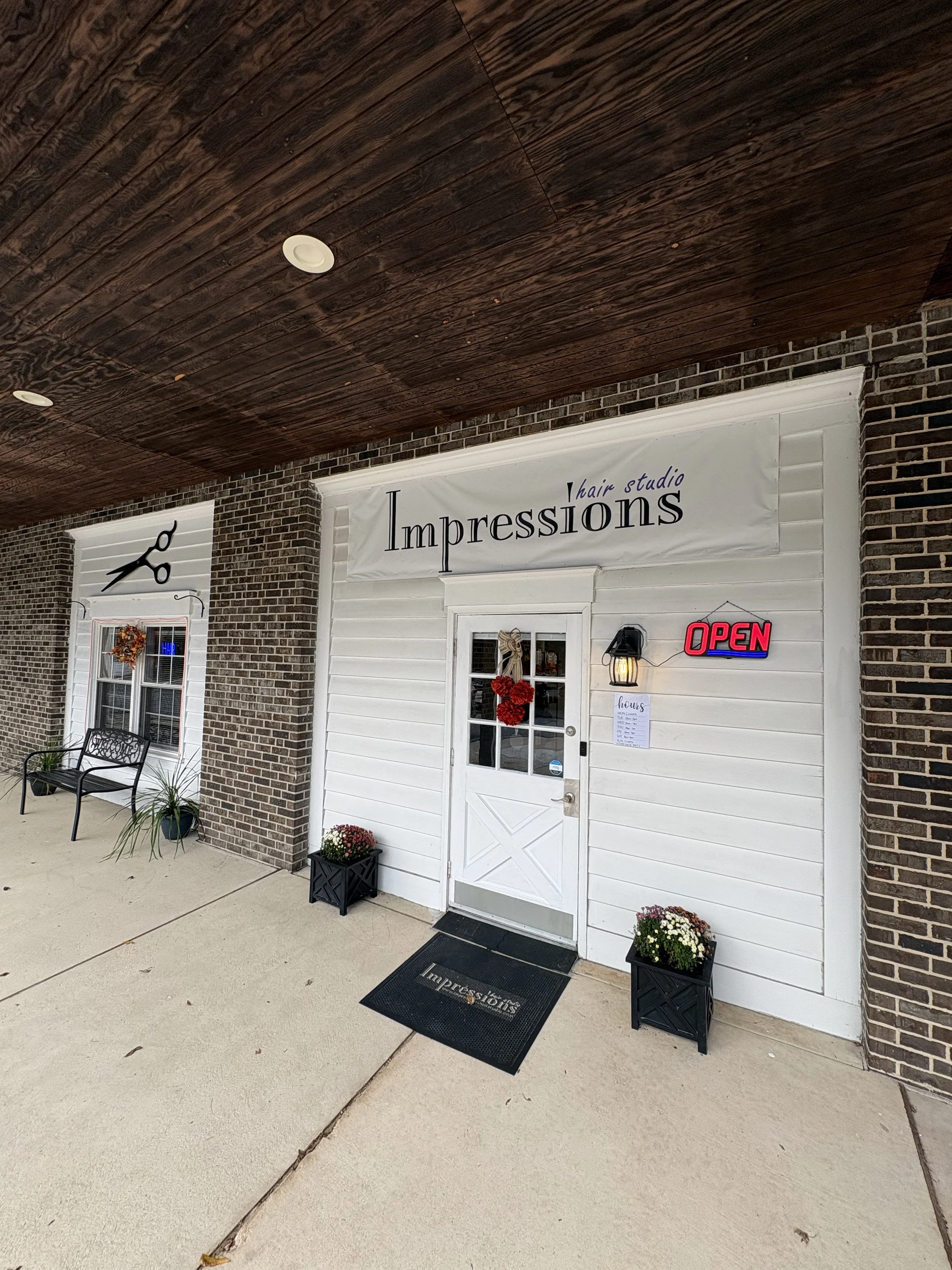 The exterior of a hair studio named Impressions, with a brick wall, white door, and an 'Open' sign. There are potted flowers, a black bench, and a doormat outside.