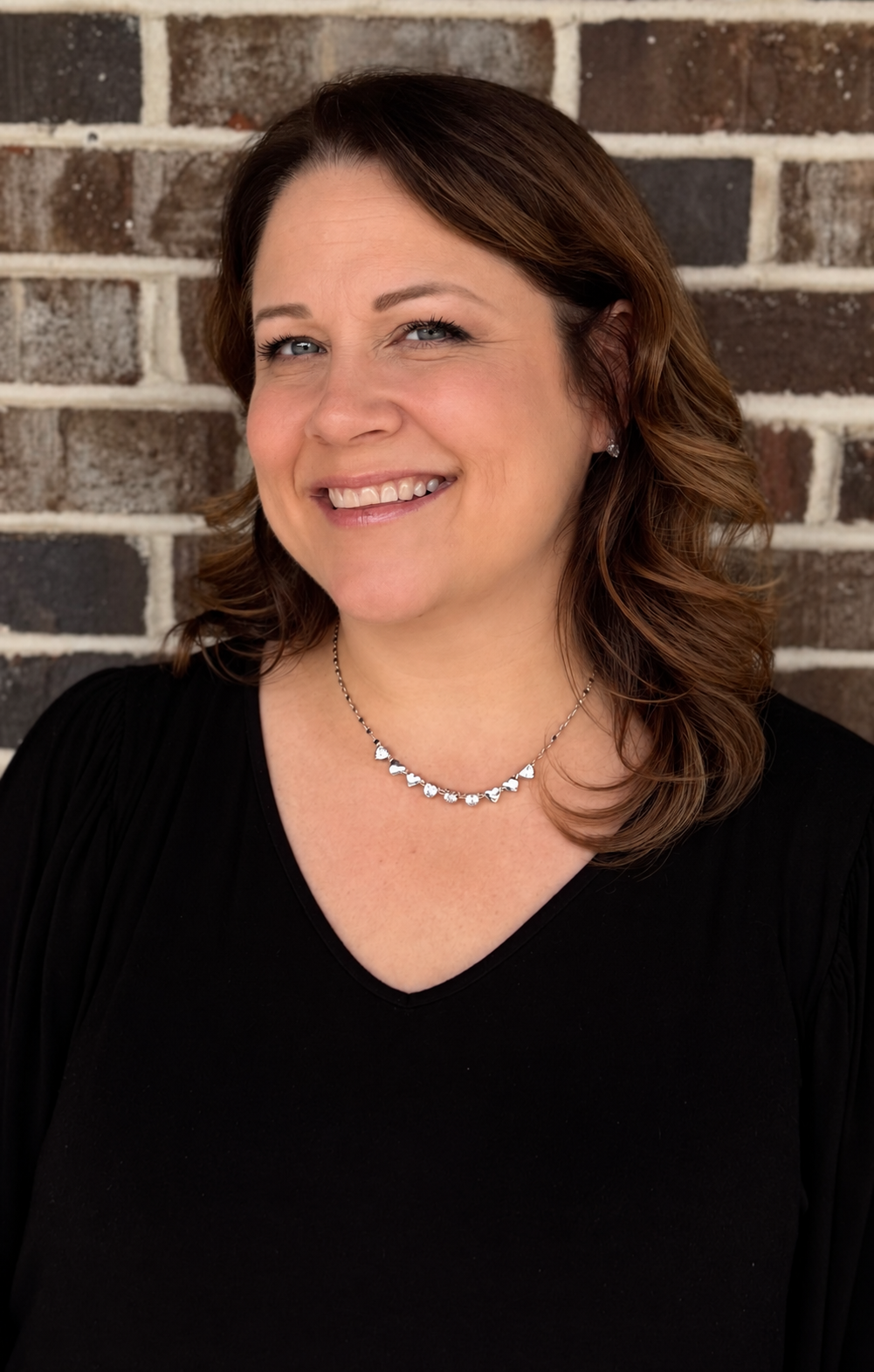 A woman with shoulder-length brown hair smiling, wearing a black top and silver necklace, standing in front of a brick wall.