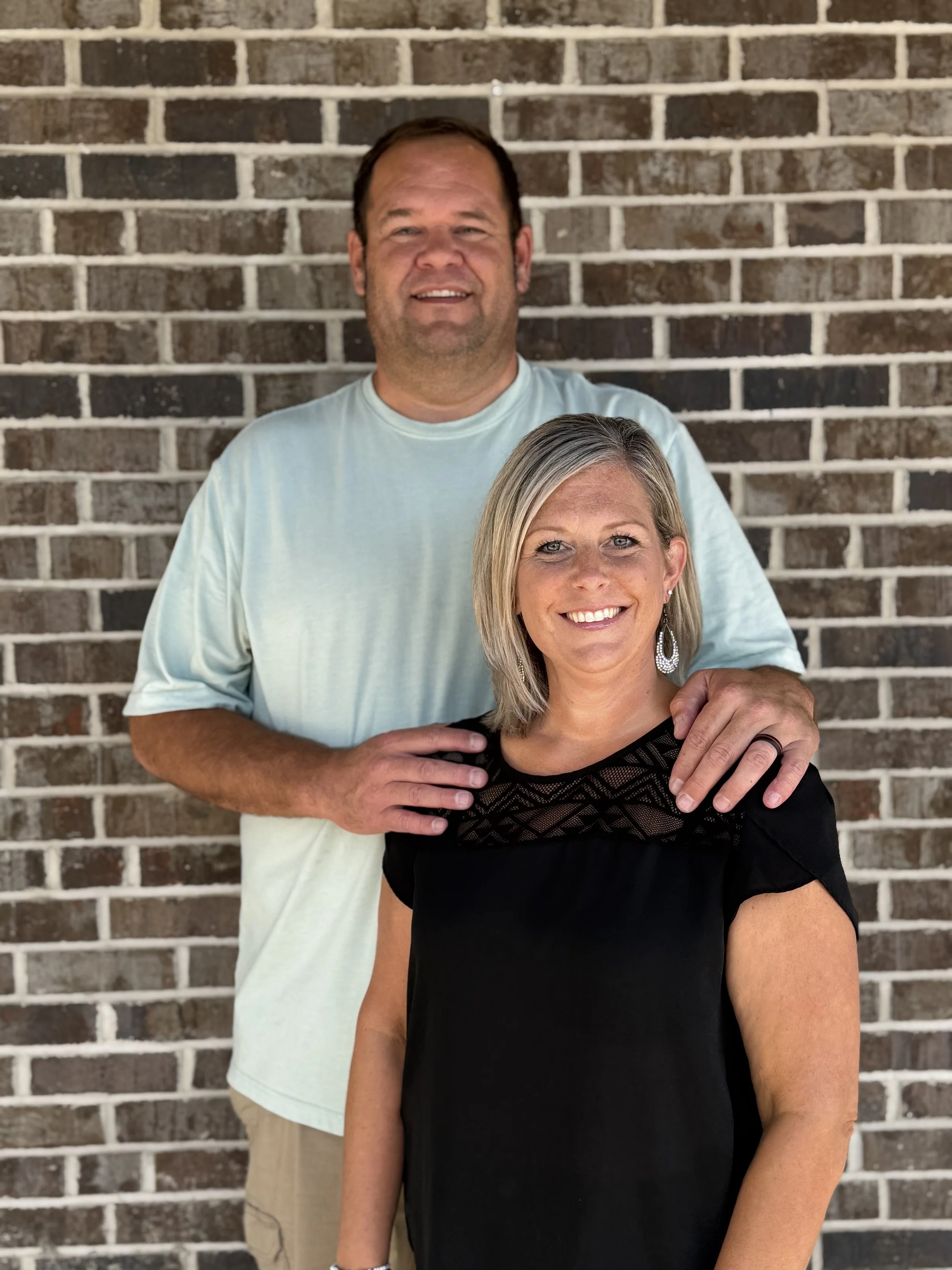 A man and a woman standing in front of a brick wall, with the man positioned behind and slightly to the left of the woman. The man has short dark hair and is wearing a light-colored T-shirt, while the woman has blonde hair styled in a shoulder-length bob and is wearing a black top with a lace detail.