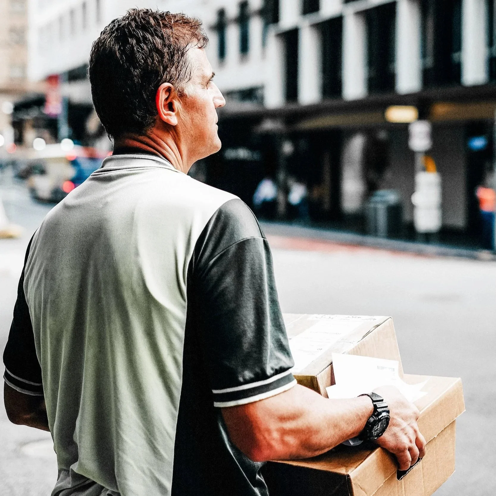 Photo of a Dependable Courier Waiting to Cross a City Street with an Armload of Packages
