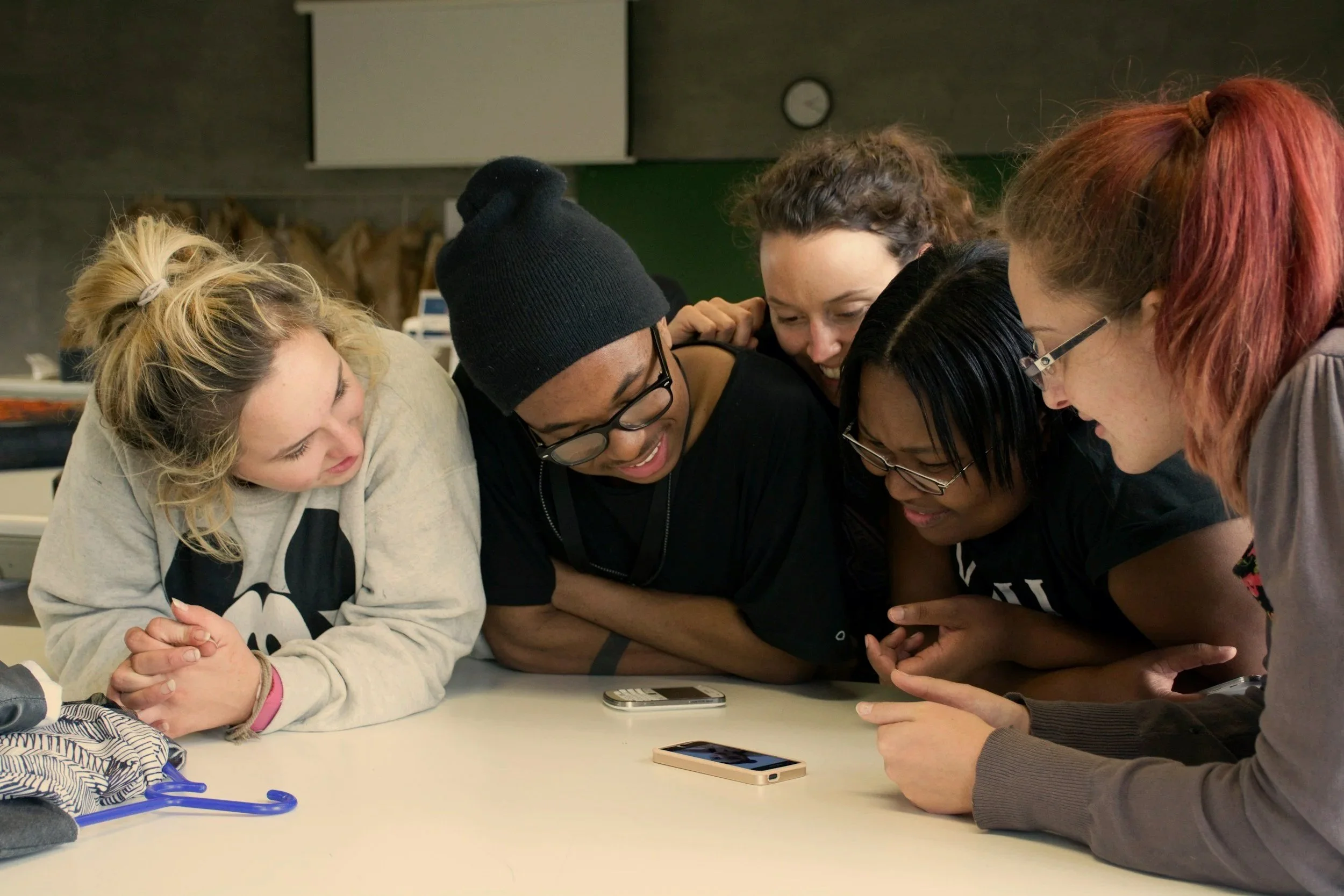 Group of five young people gathered around a table, looking at a smartphone together, with two smartphones visible on the table.