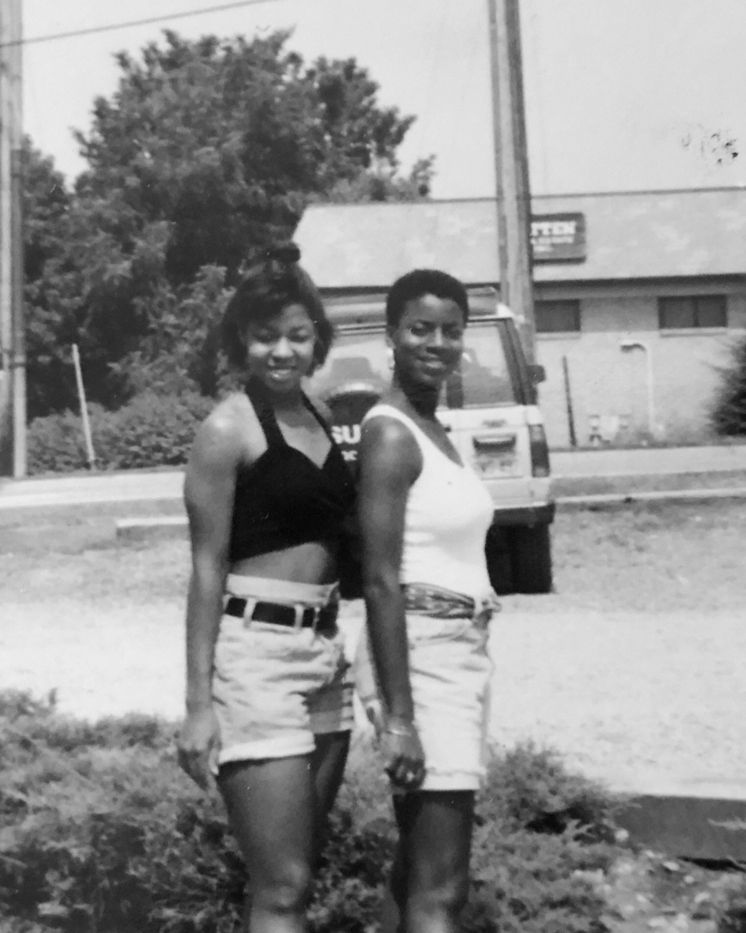 Co Founders, Naa and Verta, standing outdoors in front of a vehicle, both smiling at the camera, wearing summer clothing, with trees and a building in the background in a photo from the early 90s