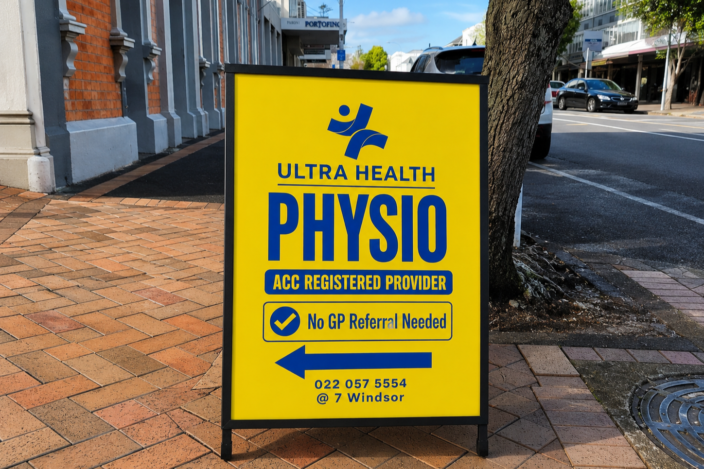 Bright yellow sidewalk sign advertising Ultra Health Physio, an ACC registered provider, with a note that no GP referral is needed, and an arrow pointing left with contact details and address.