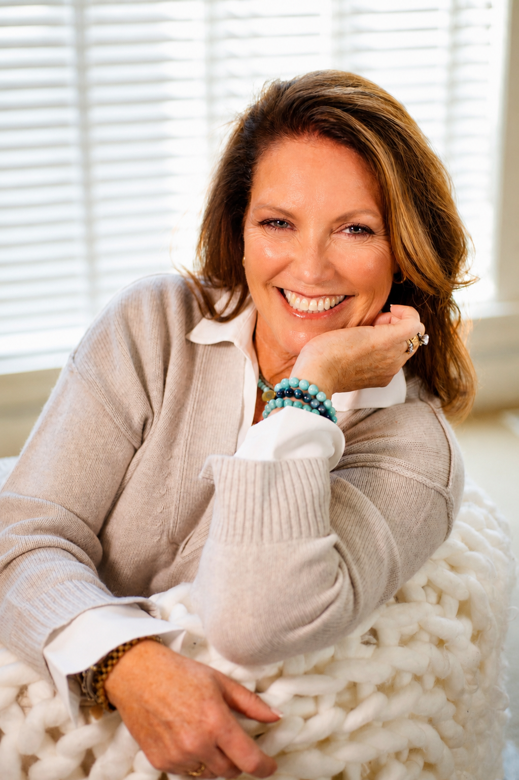 A smiling woman with shoulder-length brown hair, wearing a beige sweater over a white collared shirt, and various bracelets, resting her chin on her hand in front of a window with blinds.