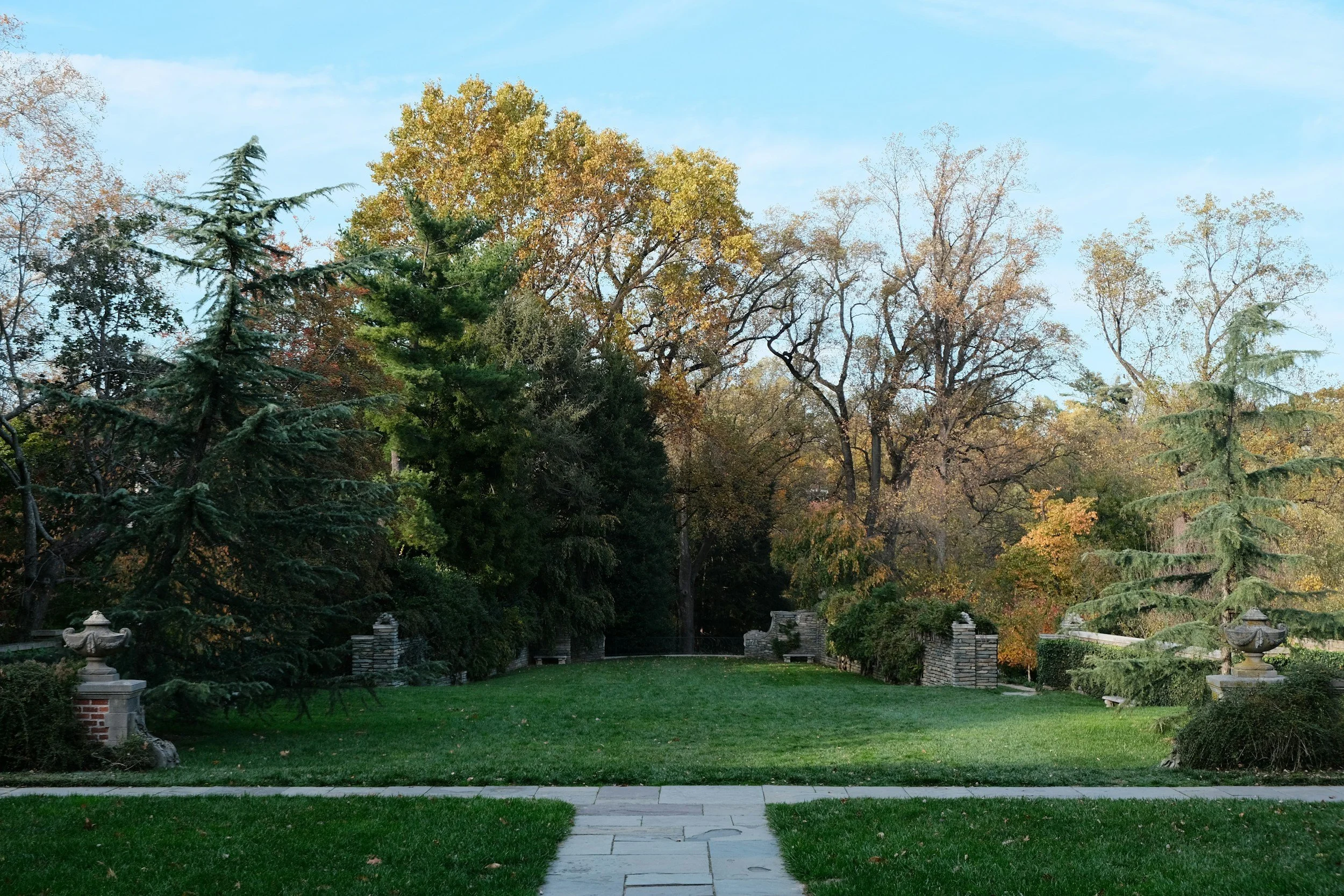 A peaceful park scene with a paved walkway, lush green grass, and tall trees with autumn-colored leaves under a clear blue sky.