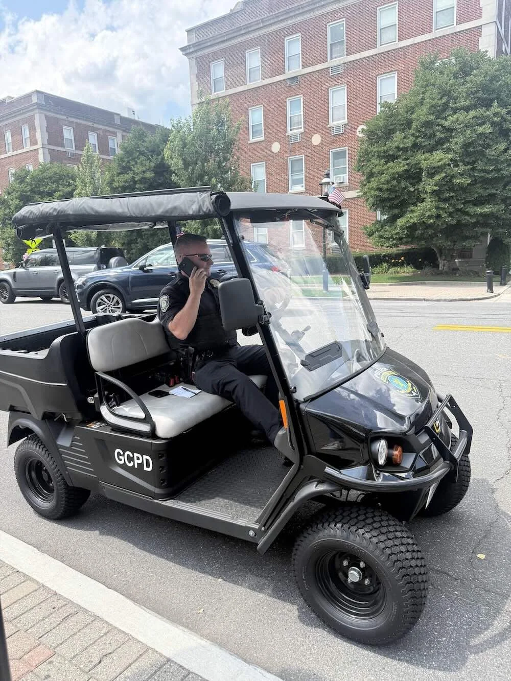 A Garden City Police Officer is sitting in a black police cart supplied by Council 1869. The golf cart is parked on the street in front of a brick building, with trees and other cars visible in the background.