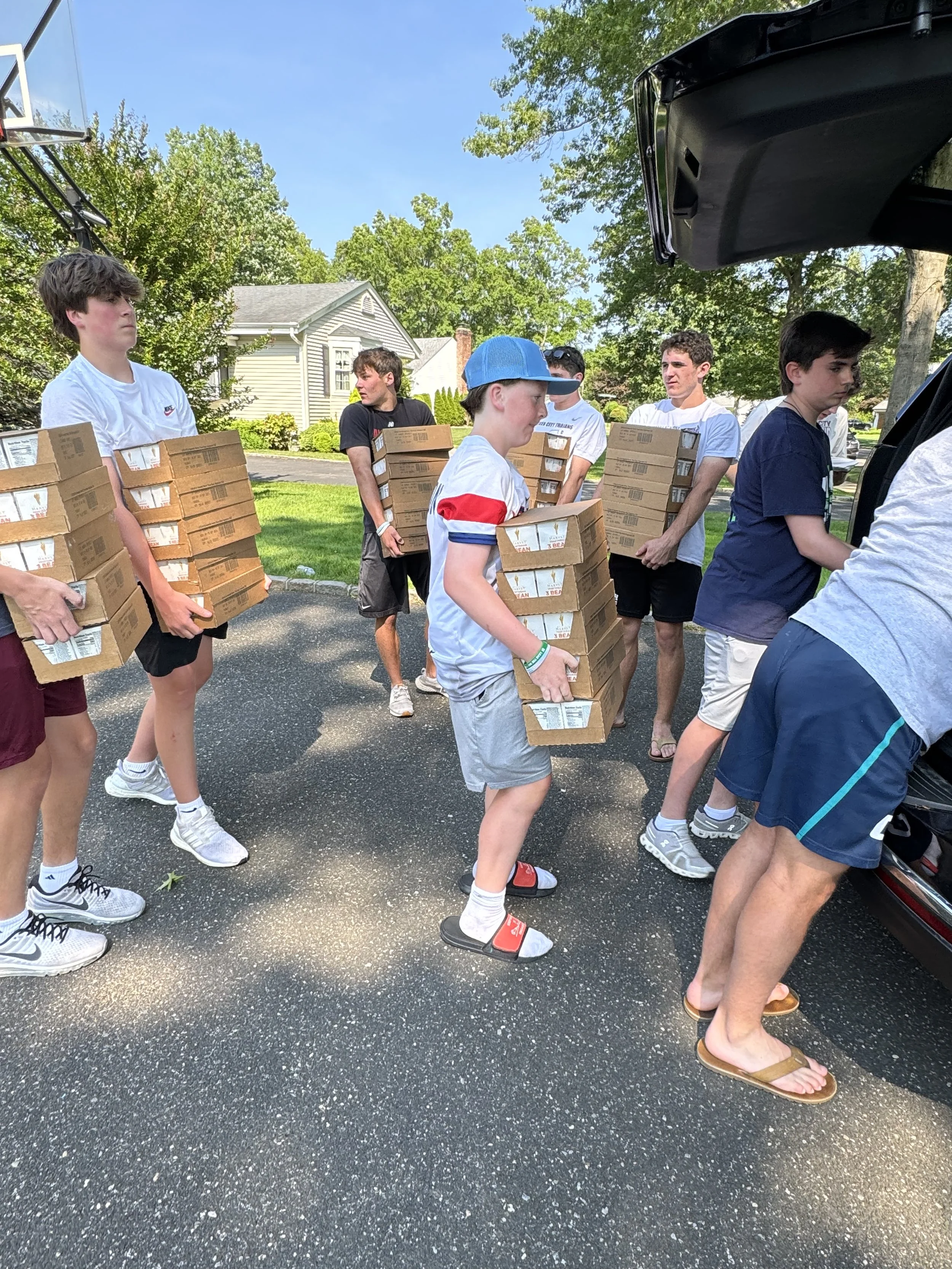 Group of young boys with standing in line, carrying pizza boxes, outdoors on a sunny day, some in shorts and t-shirts, with trees and a house in the background.