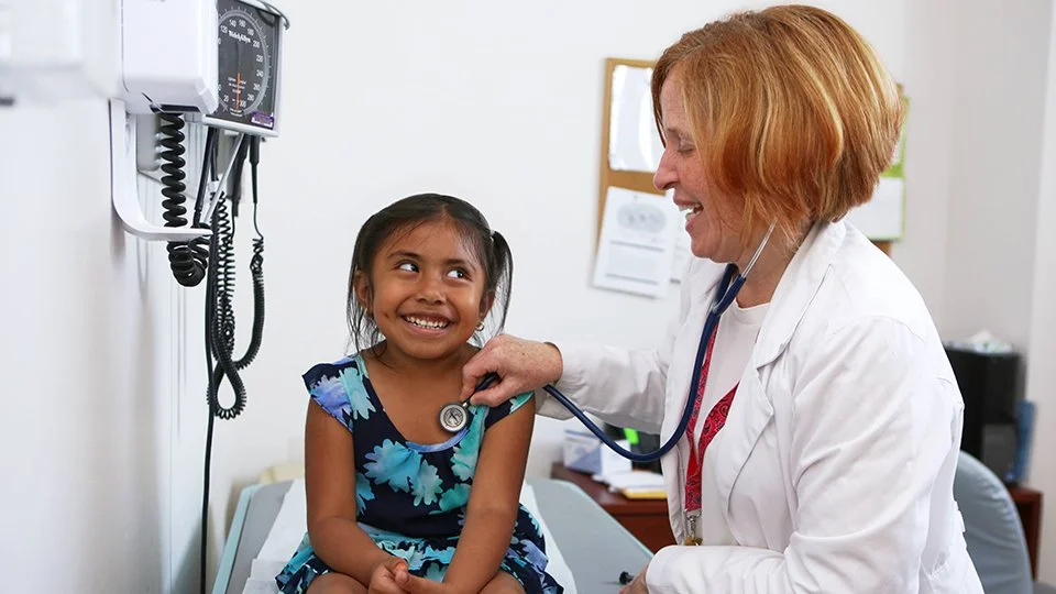 A young girl sitting on an examination table smiling while a female doctor checks her heartbeat with a stethoscope at a doctor's office.