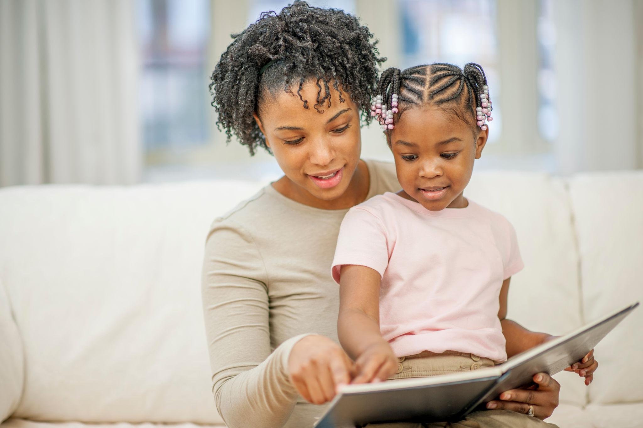 A woman and a young girl sit on a white couch, looking at a tablet together. The woman has curly hair and the girl has braided hair with pink beads.