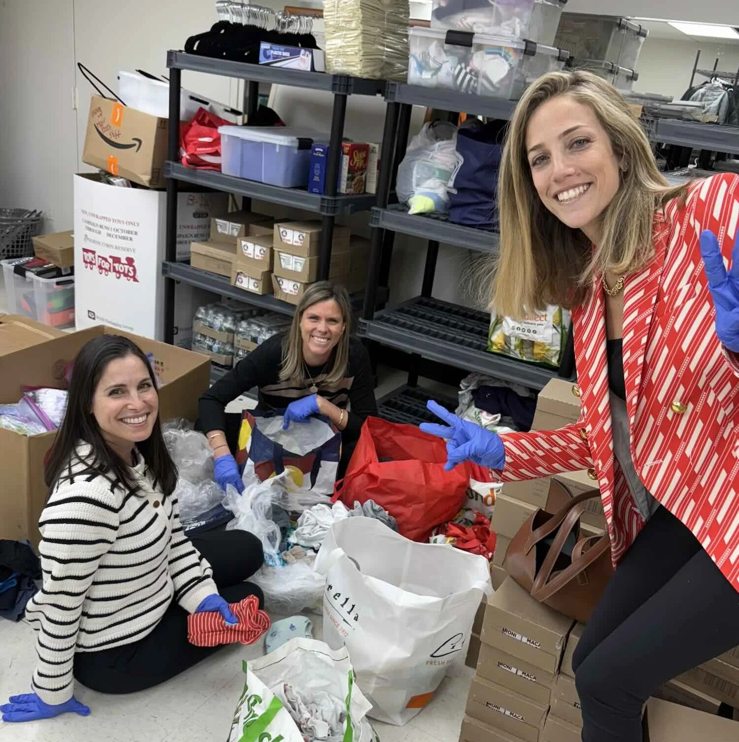 Three women wearing gloves are packing items into bags and boxes in a storage or warehouse room. They are volunteering at a council 1869 supply donation event.