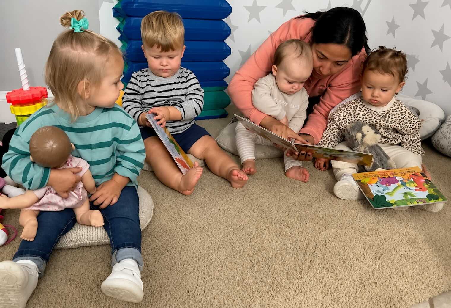 A group of young children and a woman sitting on the floor in a daycare or playroom, reading picture books together.