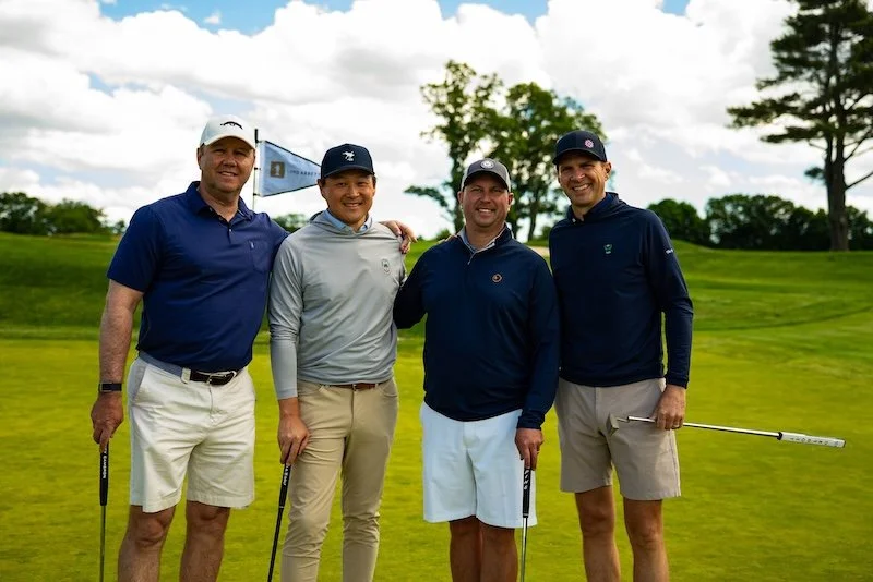 Four men standing on a golf course holding golf clubs, smiling, with trees and a cloudy sky in the background at a Council 1869 charity event