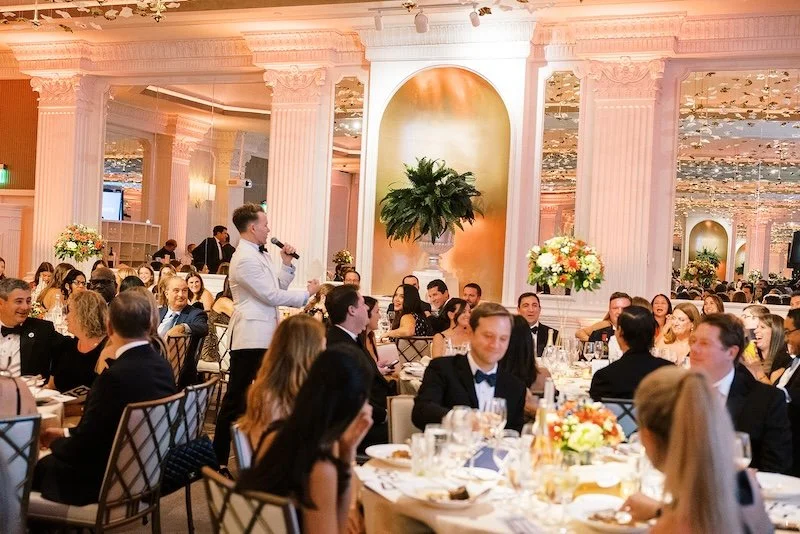 Guests seated at round tables in an elegant event hosted by Council 1869, listening to a speaker with a microphone, decorated with flowers and large plant centerpiece, ornate columns, and mirrored walls.