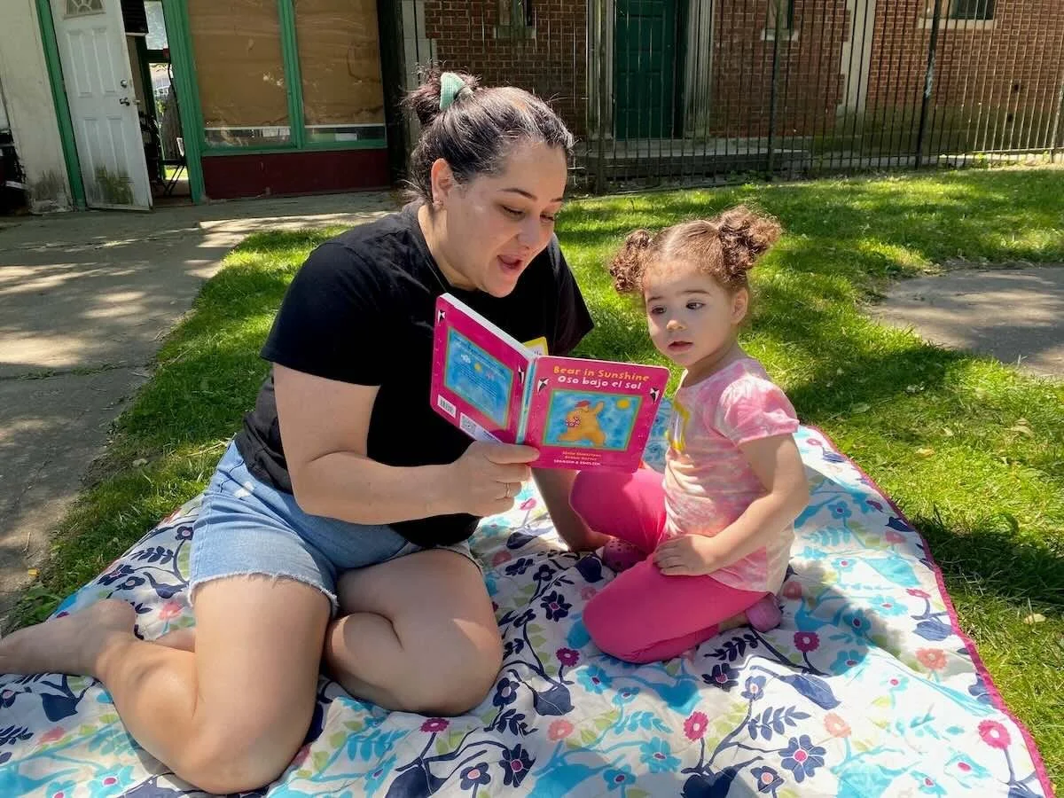 A woman and a young girl sitting on a colorful blanket outdoors, with the woman reading a children's book to the girl, during the daytime.
