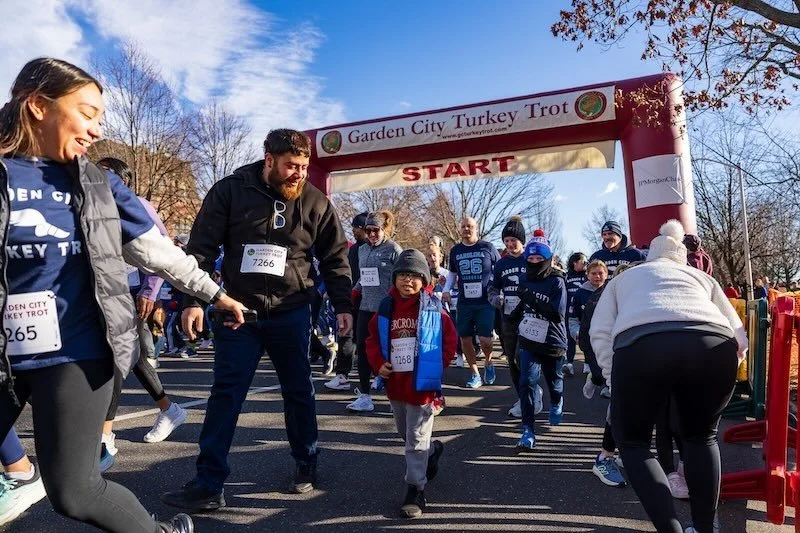 Participants at the start line of the Garden City Turkey Trot race, with some running and others walking, under a red inflatable archway with a clear blue sky overhead.