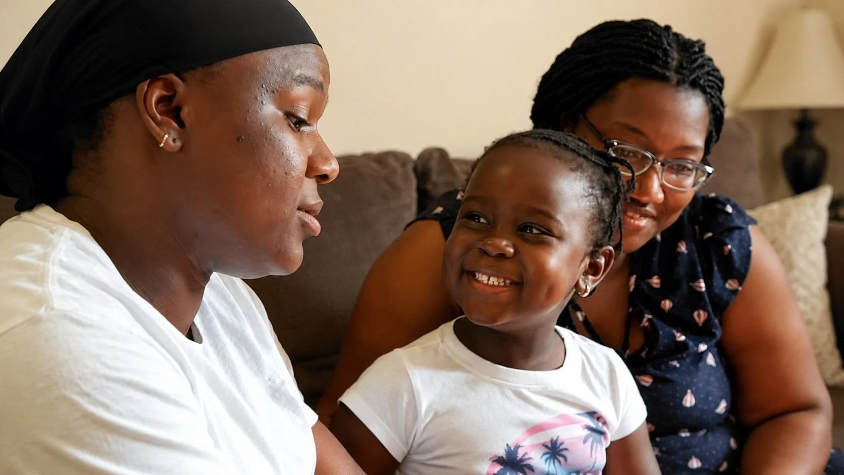 Three people from ParentChild+ sitting on a couch having a conversation. A woman in a white shirt and black headscarf, a smiling young girl with braided hair, and another woman with glasses wearing a navy blue dress with a pattern.