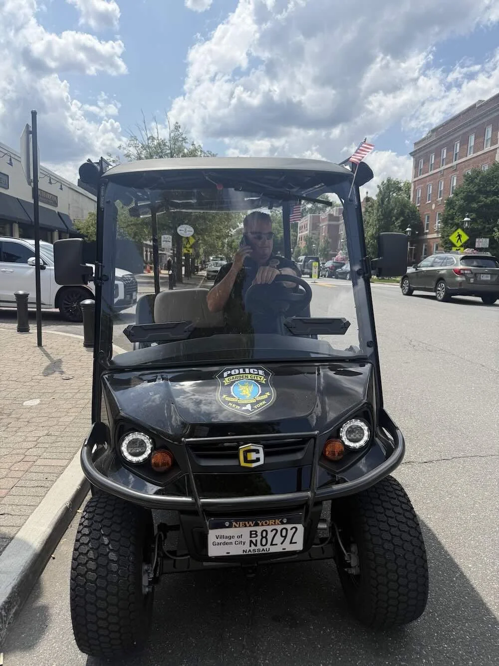 A Garden City Police Officer inside a black police golf cart supplied by Council 1869. The cart has a New York license plate, parked on a city street with buildings, cars, trees, and an American flag in the background.