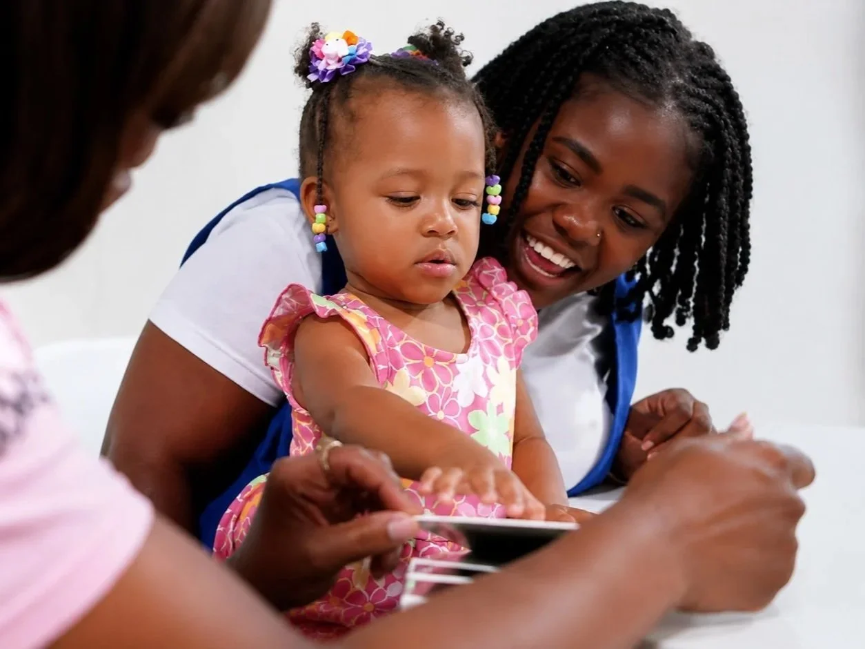 A young girl and an adult woman, likely her mother, are looking at something on a tablet device in a bright, indoor setting. The girl has colorful beads in her hair and earrings, and the woman is smiling, engaging with the girl.