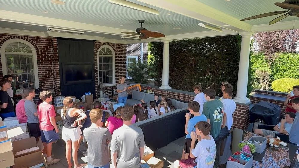 Council 1969 student impact committee meeting consisting of mostly young teenagers, gathered on a covered patio listening to a woman from council 1869 speaking. There are boxes and supplies around, and a large outdoor TV screen on the brick wall.
