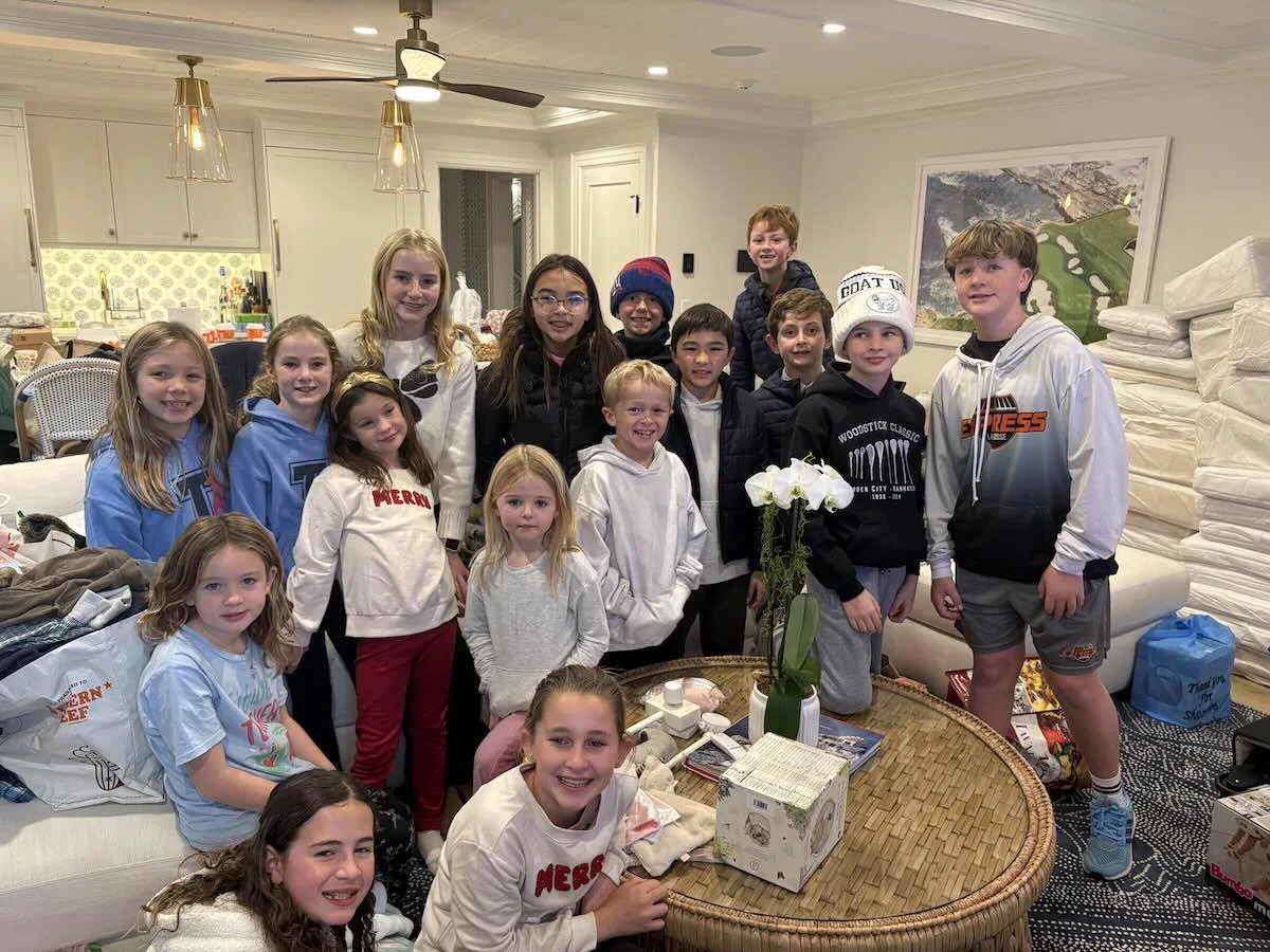 Group of children gathered in a living room, some smiling and sitting on a couch, others standing around a table with a potted orchid and various items, in a cozy indoor setting.