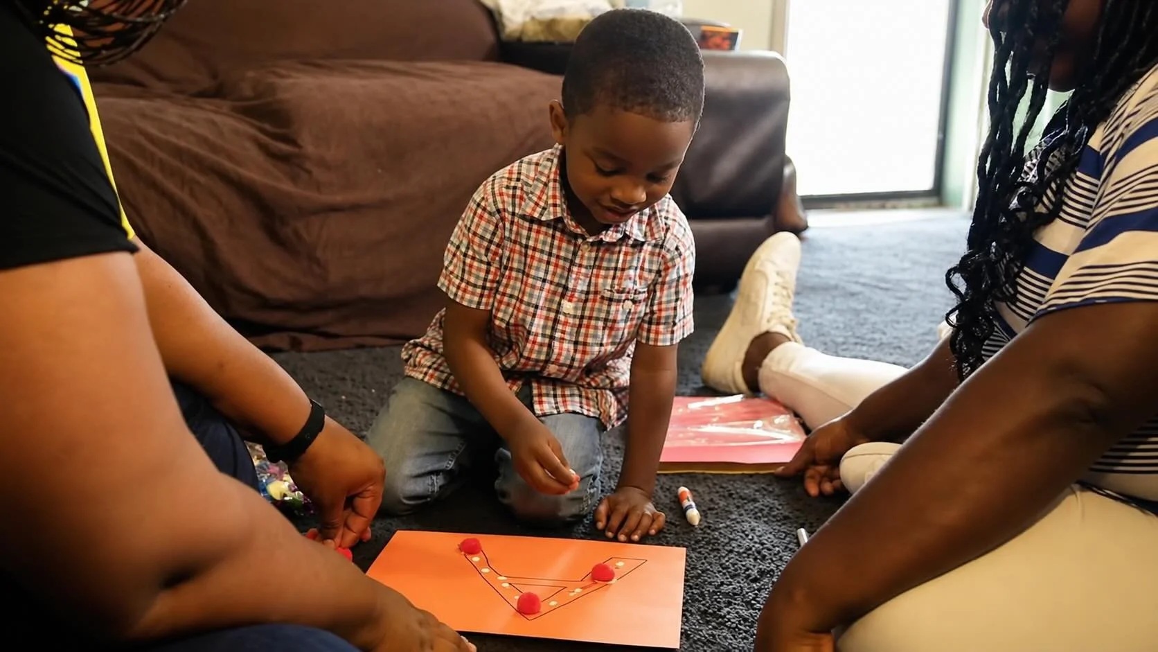 A young boy wearing a plaid shirt and jeans is sitting on the floor, playing a game with two adults who are also sitting on the floor. This is an In Home Eductational Visit by ParentChild+, supported by Council 1869