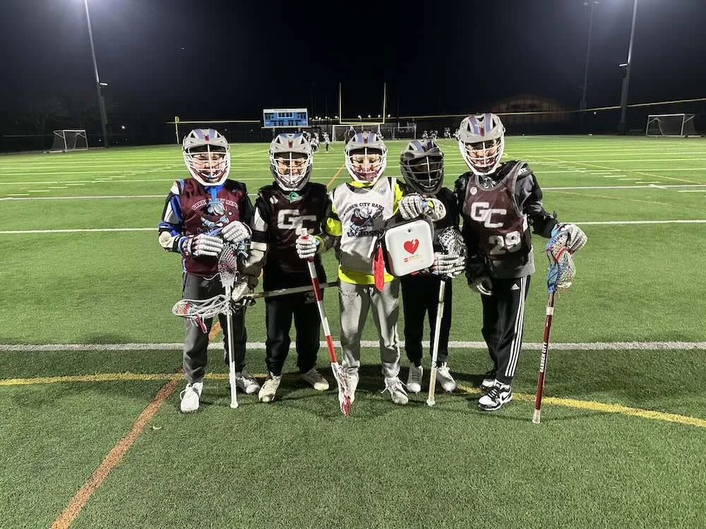 Group of five young garden city lacrosse players wearing helmets and gear on a football field at night, holding lacrosse sticks and the new AED device purchased by Council 1869