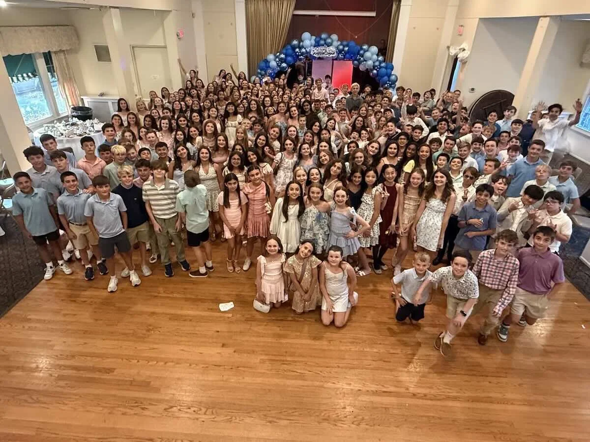 Large group of children and teenagers at a party or celebration, standing on a wooden dance floor in a decorated hall with balloon arch and curtains. This is the Garden City Autism Ball that Council 1869 supports.