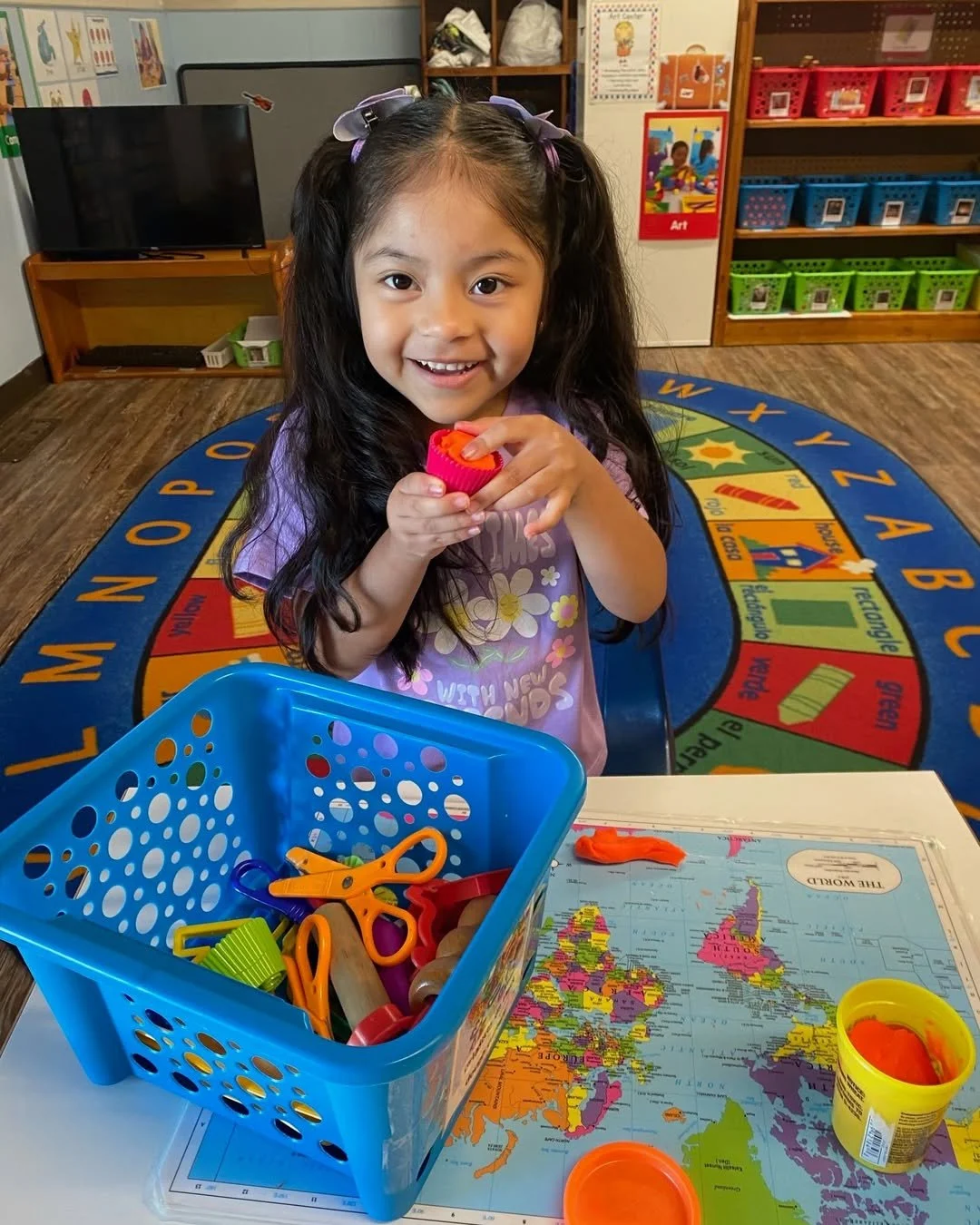 A young girl with long dark hair, wearing a purple shirt with flowers, is sitting at a table in a classroom. She is smiling and holding a small orange playdough cup. In front of her are some orange playdough, a toy pink knife, and a yellow container 