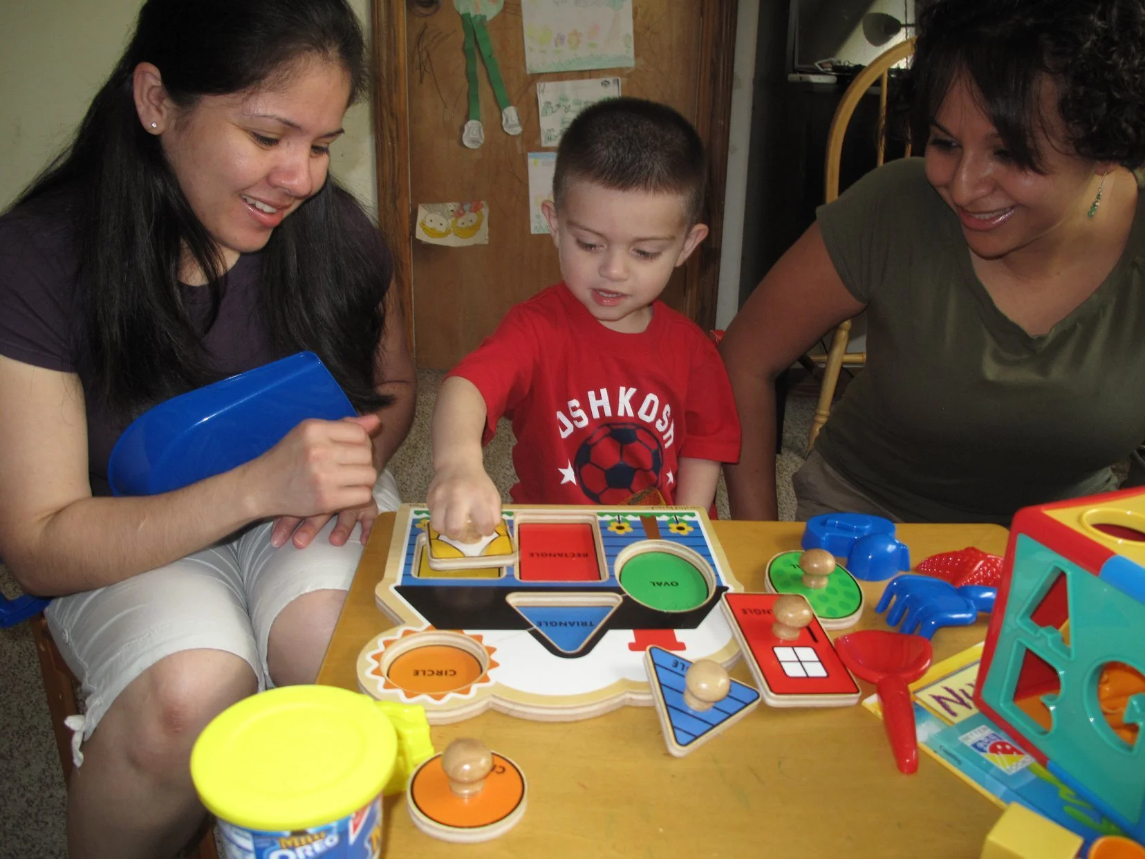 A young boy playing a board game with two women beside him, sitting at a table filled with various toy game pieces and components.