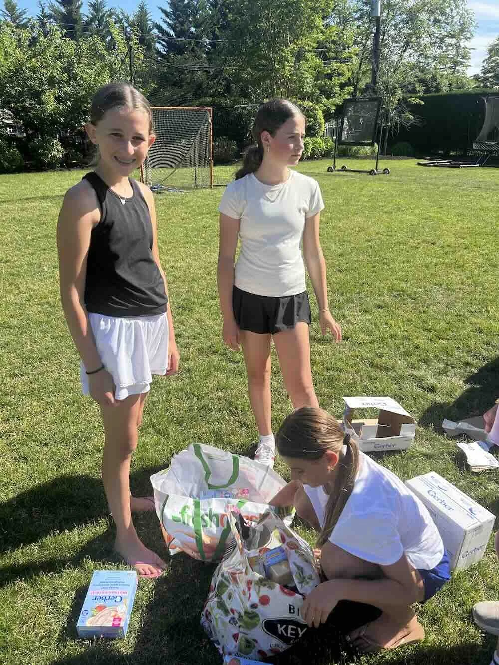 Three girls in sportswear are outdoors on a grassy area. One girl in a black top and white shorts is smiling at the camera. A second girl in a white shirt and black shorts looks to the side. The third girl is kneeling, searching through a bag on the ground, surrounded by grocery boxes and bags. In the background, there is a tennis net, trees, and sports equipment.