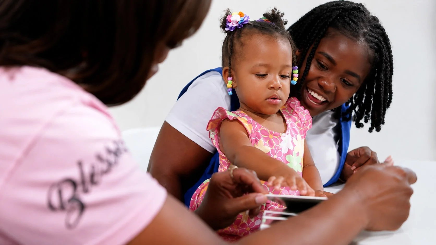 A woman, a young girl, and a healthcare provider sitting together, looking at a tablet. The young girl has colorful bead earrings and hair, and is wearing a pink dress with floral patterns. The woman and healthcare provider are smiling as they look a