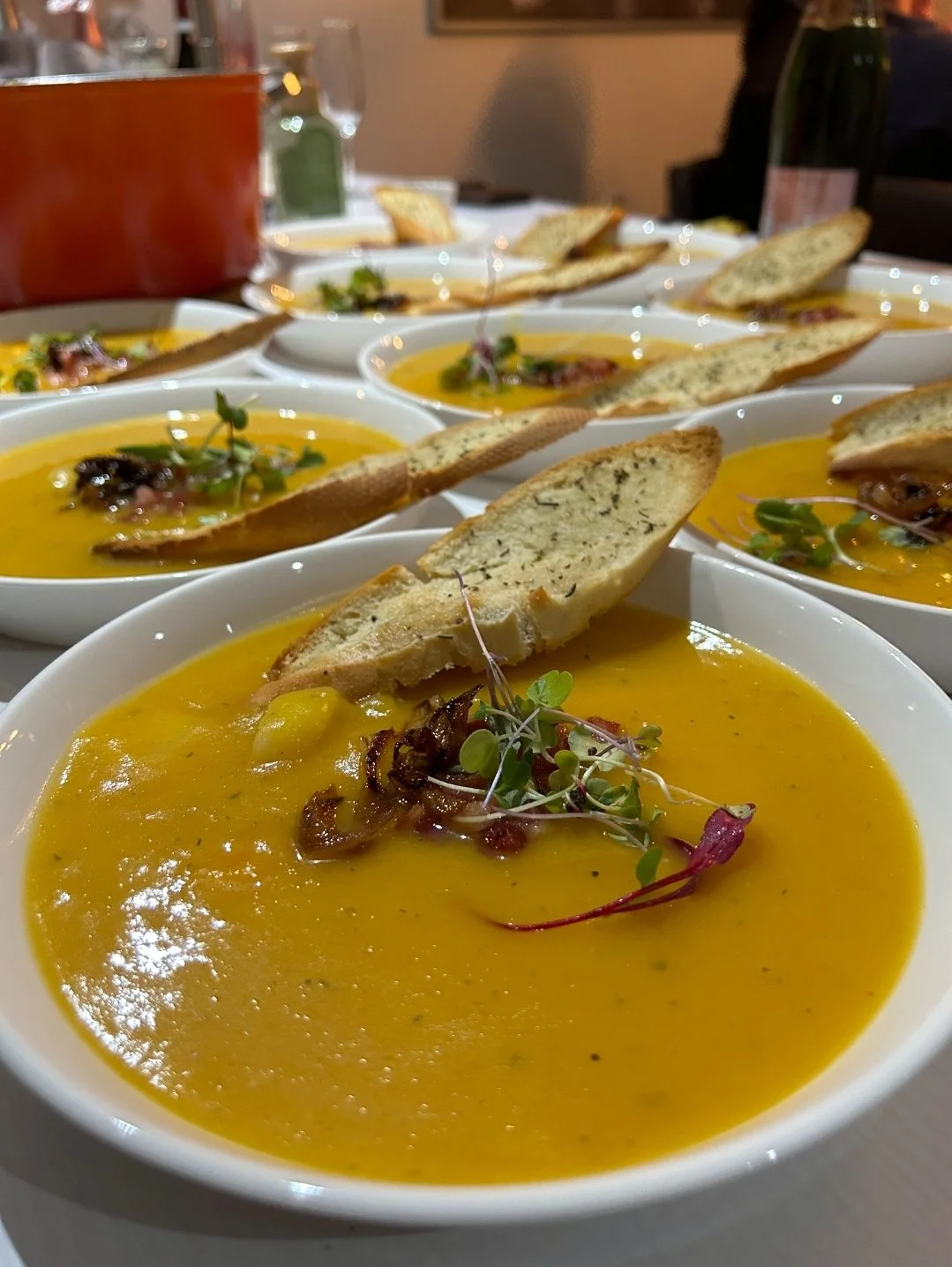 Several bowls of butternut squash soup garnished with microgreens, crispy bacon, and toasted bread slices on a dining table.